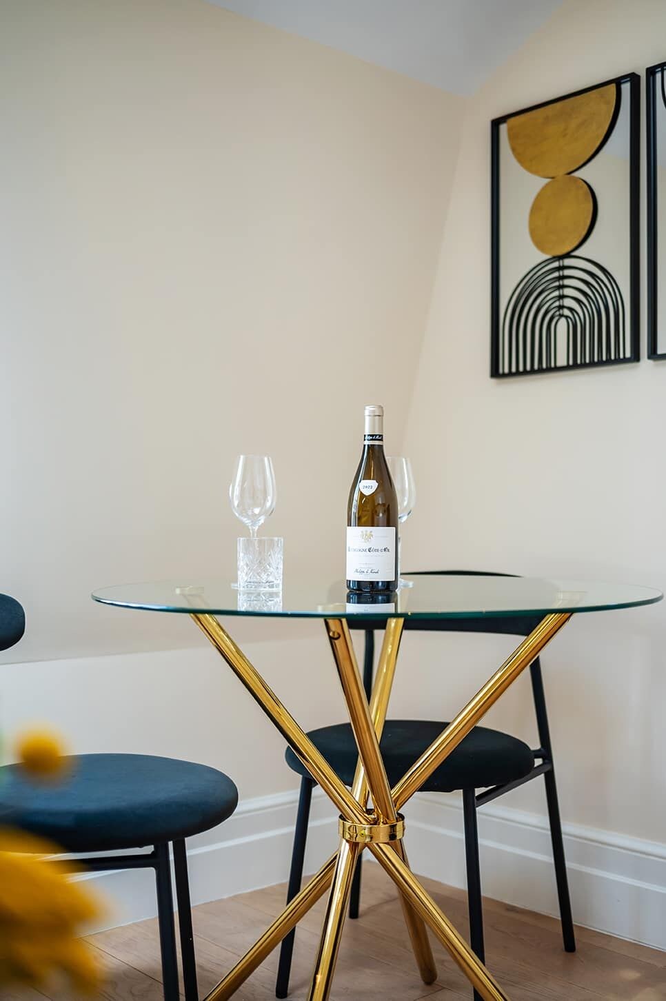 En matplats med ett runt bord i glas, guldfärgade ben och svarta stolar. En vinflaska och två vinglas står på bordet.