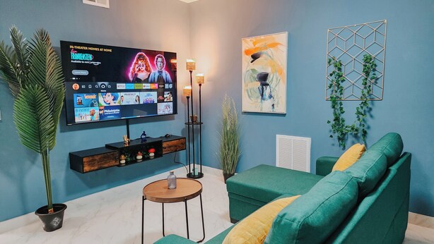 A living room with a flat-screen TV, green sofas, a coffee table, and decorative plants.