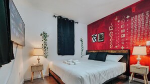 A bedroom with a bed, two bedside tables, a wall-mounted TV, and a red patterned wall.
