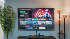 A flat-screen TV mounted on a wall displaying a streaming service interface with a movie titled "In Theater Movies at Home" and a "Live" section.