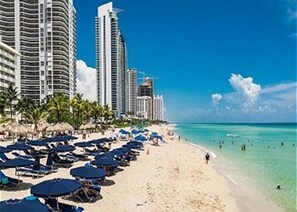 A beachside hotel with multiple high-rise buildings, a sandy beach, and numerous umbrellas. - G - Walk to beach!  Beautiful Pool and Tennis Courts (Sunny Isle)