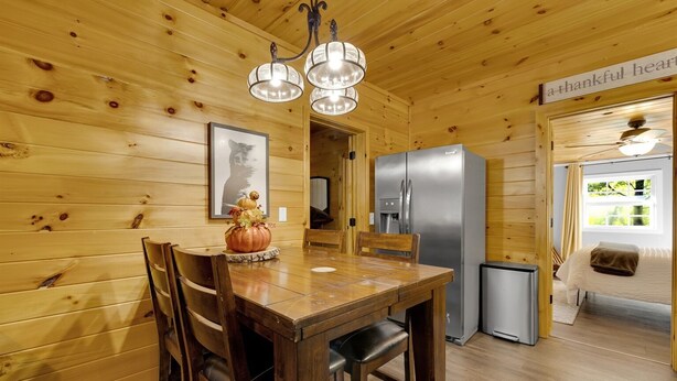 A wooden interior with a dining table, chairs, a chandelier, a refrigerator, and a bed with a window view.