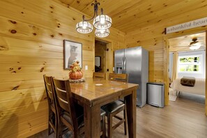 Un interior de madera con una mesa de comedor, sillas, una araña, un refrigerador y una cama con vistas al exterior.