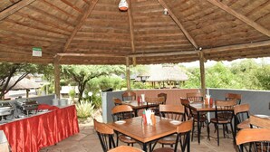 Un área de comedor al aire libre con mesas y sillas de madera, una barra de buffet y una estructura de techo de paja al fondo.