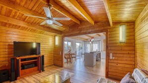 A wooden interior with a TV, glass coffee table, ceiling fan, and a kitchen area visible through an open door.
