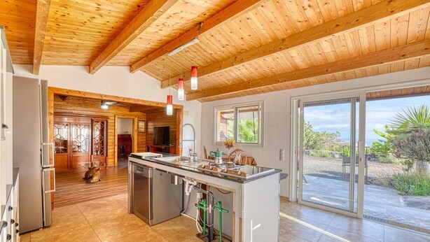 A modern kitchen with a central island, wooden ceiling beams, and a view of the outdoors.