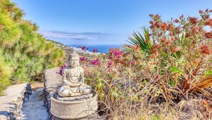 A Buddha statue on a stone pedestal by a coastal path with vegetation and a clear sky.