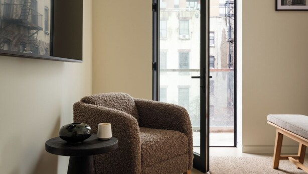Un salon moderne avec un fauteuil marron, une petite table noire sur laquelle se trouvent un bol et une tasse, un banc en bois et une grande fenêtre donnant sur la ville.