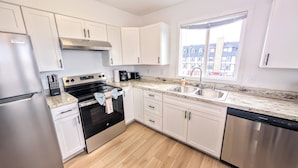 A modern kitchen with white cabinets, stainless steel appliances, a double sink, and a window providing a view of buildings outside. - Brand New Large 2Bd/1Ba Apartment - Near Downtown, King Bed! (Anchorage)