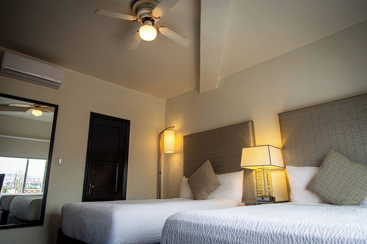 Habitación de hotel con dos camas, ventilador de techo, unidad de aire acondicionado, mesita de noche con lámpara y un espejo grande.