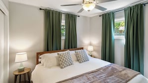 Un lit bien fait, avec une tête de lit en bois, deux coussins à motifs et un plaid marron. On y trouve une petite table de chevet avec une lampe, et la pièce est dotée de rideaux verts et d’un ventilateur de plafond.
