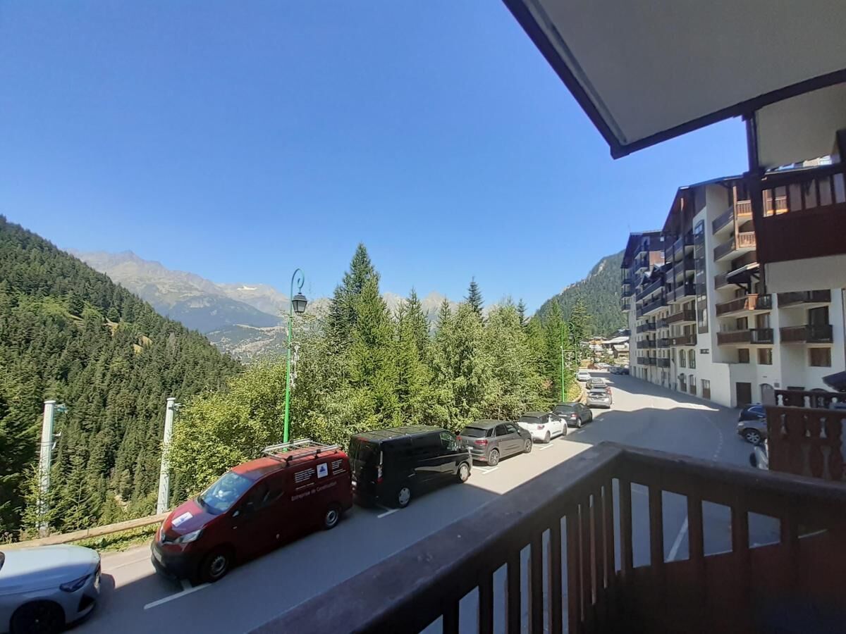 Une route de montagne bordée de véhicules garés, dont un fourgon rouge et un camion noir, offrant une vue sur une vallée boisée et des montagnes lointaines.