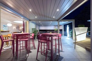Un área de comedor al aire libre con sillas y mesas de metal rojo, techo de madera y vista a la cocina y al jardín.