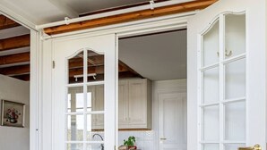 A white door leading to a kitchen with wooden beams, a rug, and a hanging light fixture.