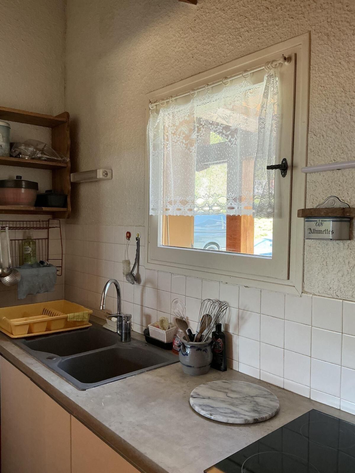 Una cucina con un lavello, una finestra con tende in pizzo, uno scaffale con oggetti da cucina e un piano di lavoro in marmo.