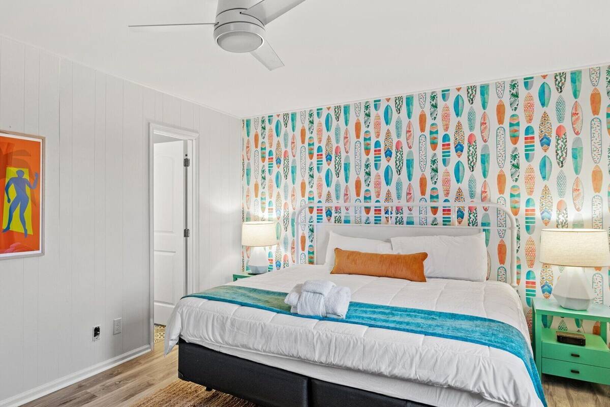A bedroom with a bed, a ceiling fan, a colorful wall with surfboard patterns, and a framed artwork on the wall.