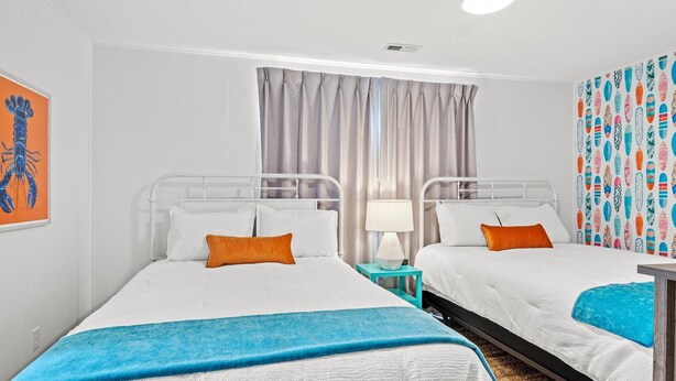 Une chambre d’hôtel avec deux lits, une fenêtre avec des rideaux, une décoration murale colorée et une petite table avec une lampe.