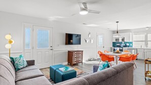 A modern living room with a grey sofa, a coffee table, and orange chairs. There is a kitchen area with white cabinets and a blue backsplash. - Beach Nest - R R Beach Club (North Myrtle Beach)