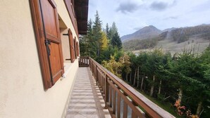 A balcony with a tiled floor and wooden railing, overlooking a mountainous landscape with trees and a clear sky.