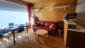 A cozy living room with a wooden ceiling, a red leather sofa, a dining table with chairs, a small round table, and a kitchenette area with a microwave and coffee maker. - 300m from ski slopes, Ground floor, view parking, terrace, parking, tv, ski locker, 29m² (Montgenèvre)