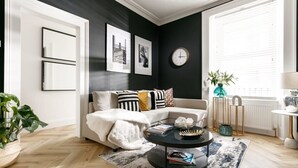 A modern living room with a white sofa, black and white cushions, a black coffee table, and a large window with blinds. - The Luxe Experience House | Central Plymouth | Sleeps 9 (Plymouth)