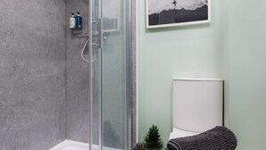 Un baño con ducha envuelta en vidrio, inodoro con reposacabezas decorativo y un cuadro en la pared.