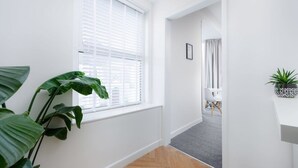Een gang met een witte muur, een raam met jaloezieën, een plant in pot en een grijs tapijt.