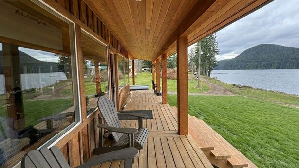 Eine Holzterrasse mit Sitzgelegenheiten im Freien, großen Fenstern und Blick auf einen See und Berge.