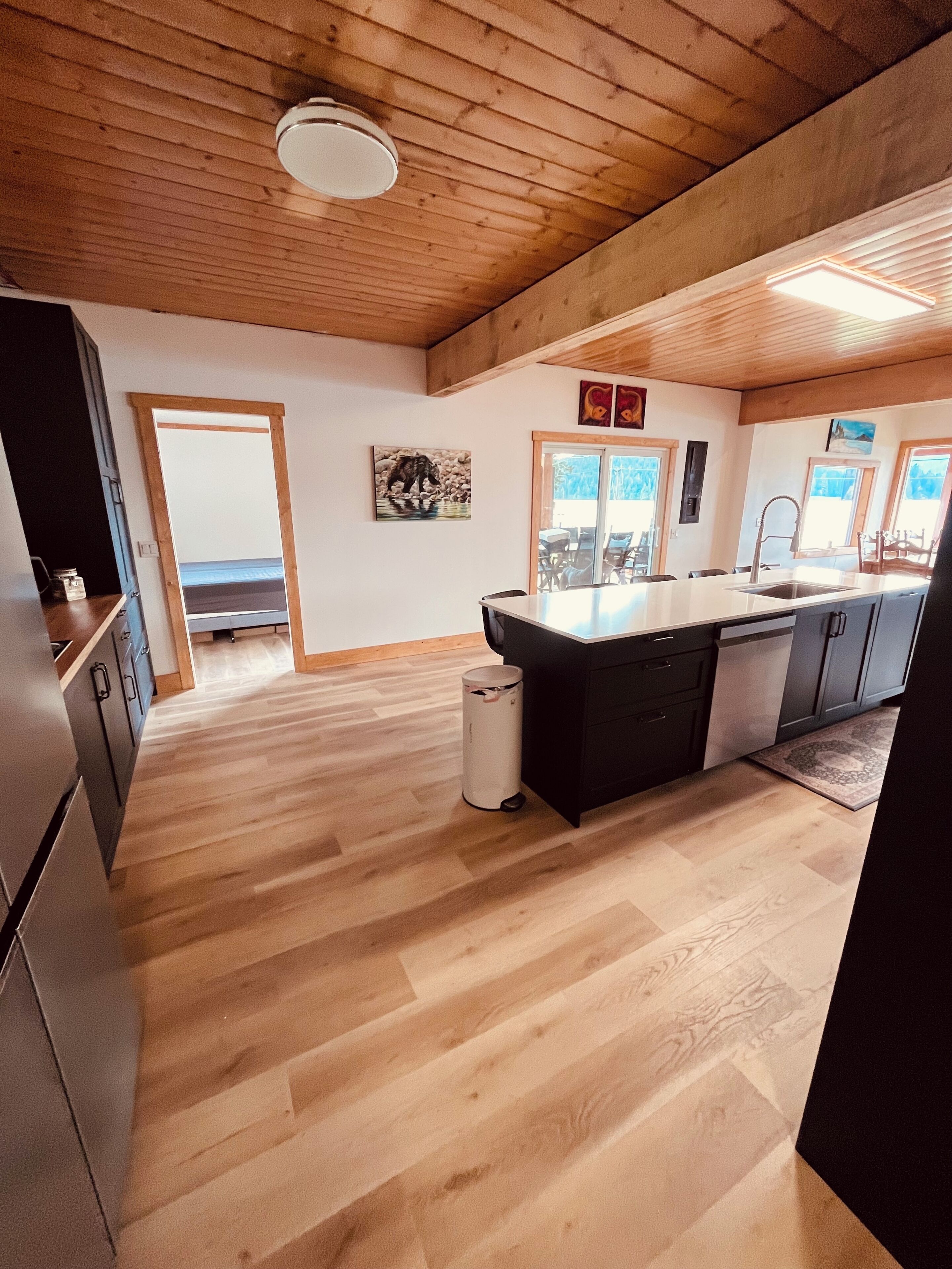 Une cuisine spacieuse avec un plancher en bois, un îlot central et une vue sur l’extérieur, visible à travers la porte.