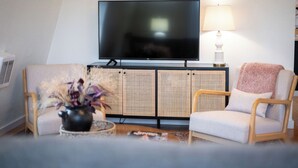 Una sala de estar con una televisión de pantalla plana, un mueble de ratán, dos sillas, una mesa con un jarrón y una lámpara.
