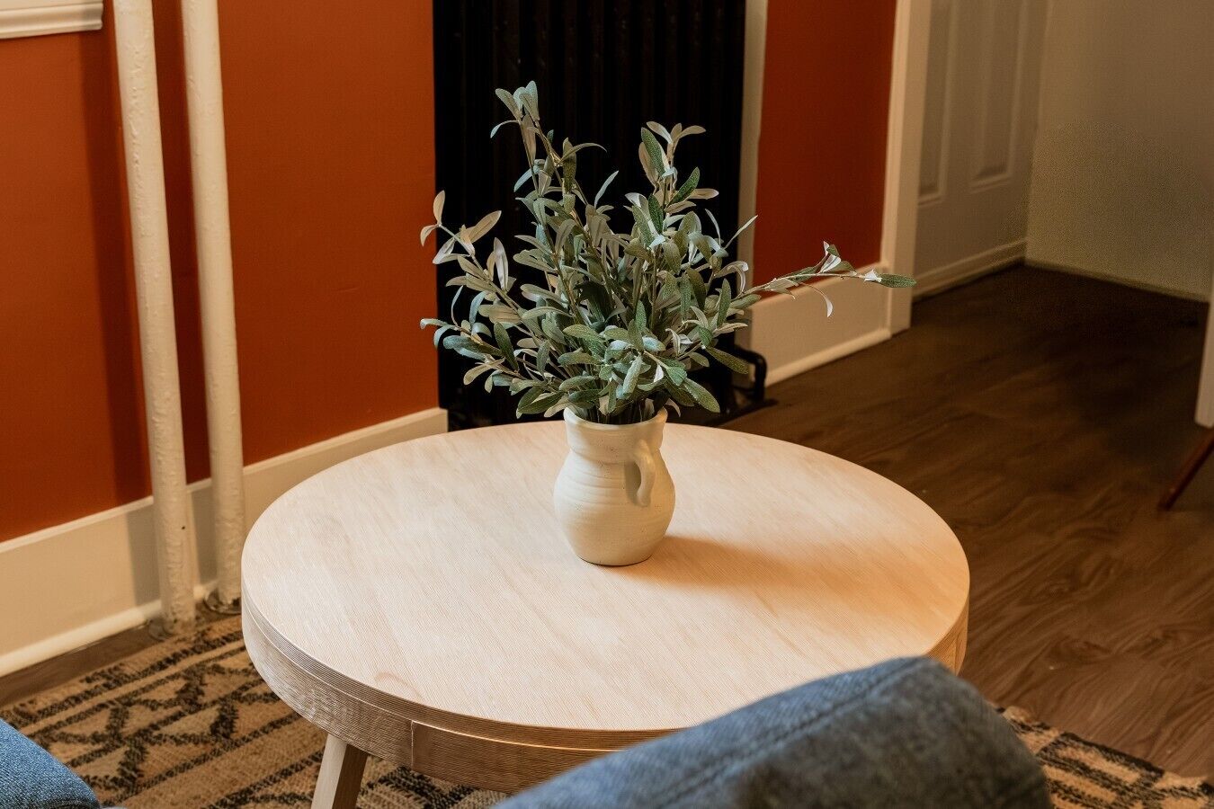 Et rundt træbord med en hvid vase med en plante, placeret i et rum med orange vægge og et mønstret tæppe.