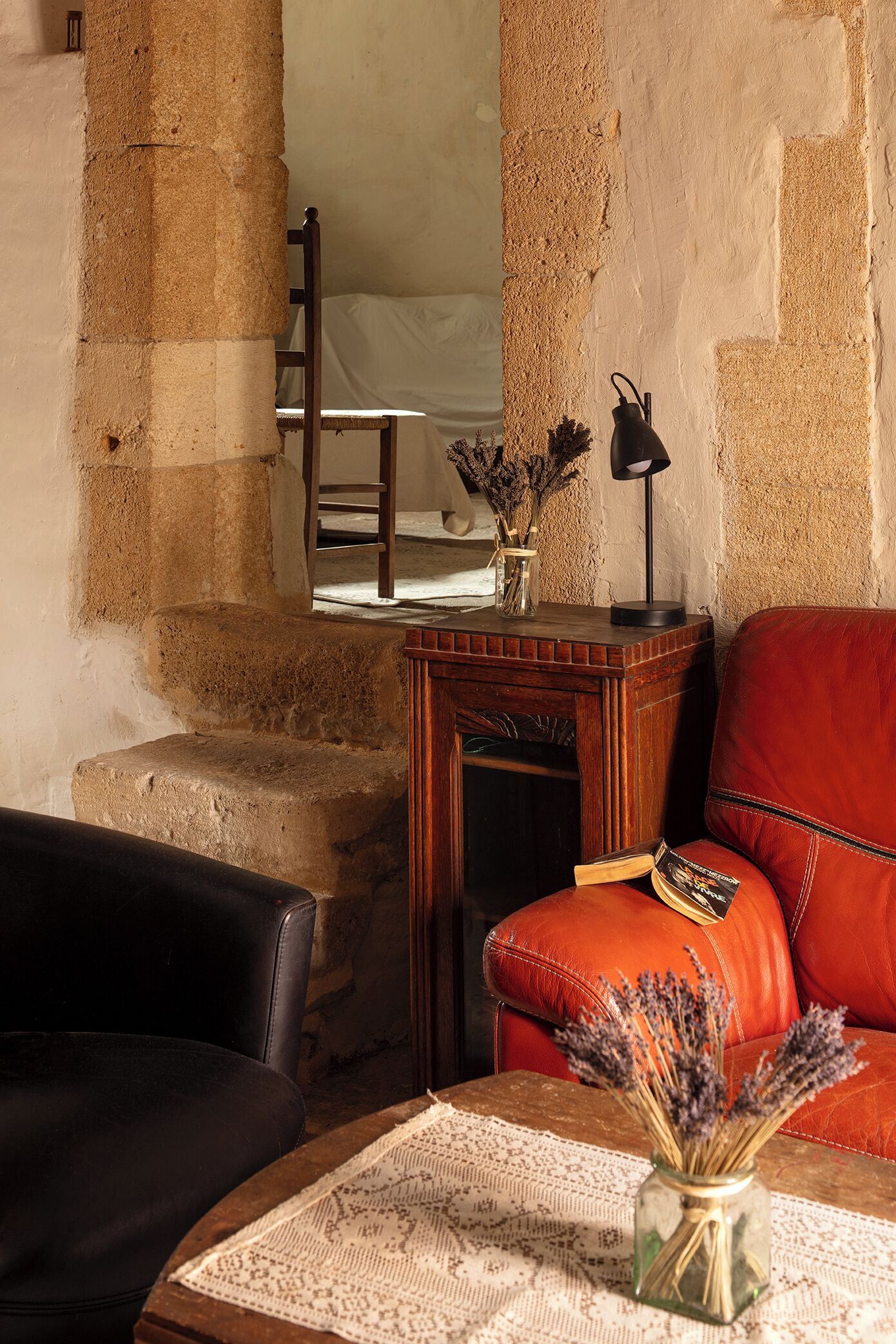 Une pièce avec un mur en pierre, une table en bois sur laquelle est posé un vase de fleurs séchées, un fauteuil en cuir rouge et un canapé en cuir noir.