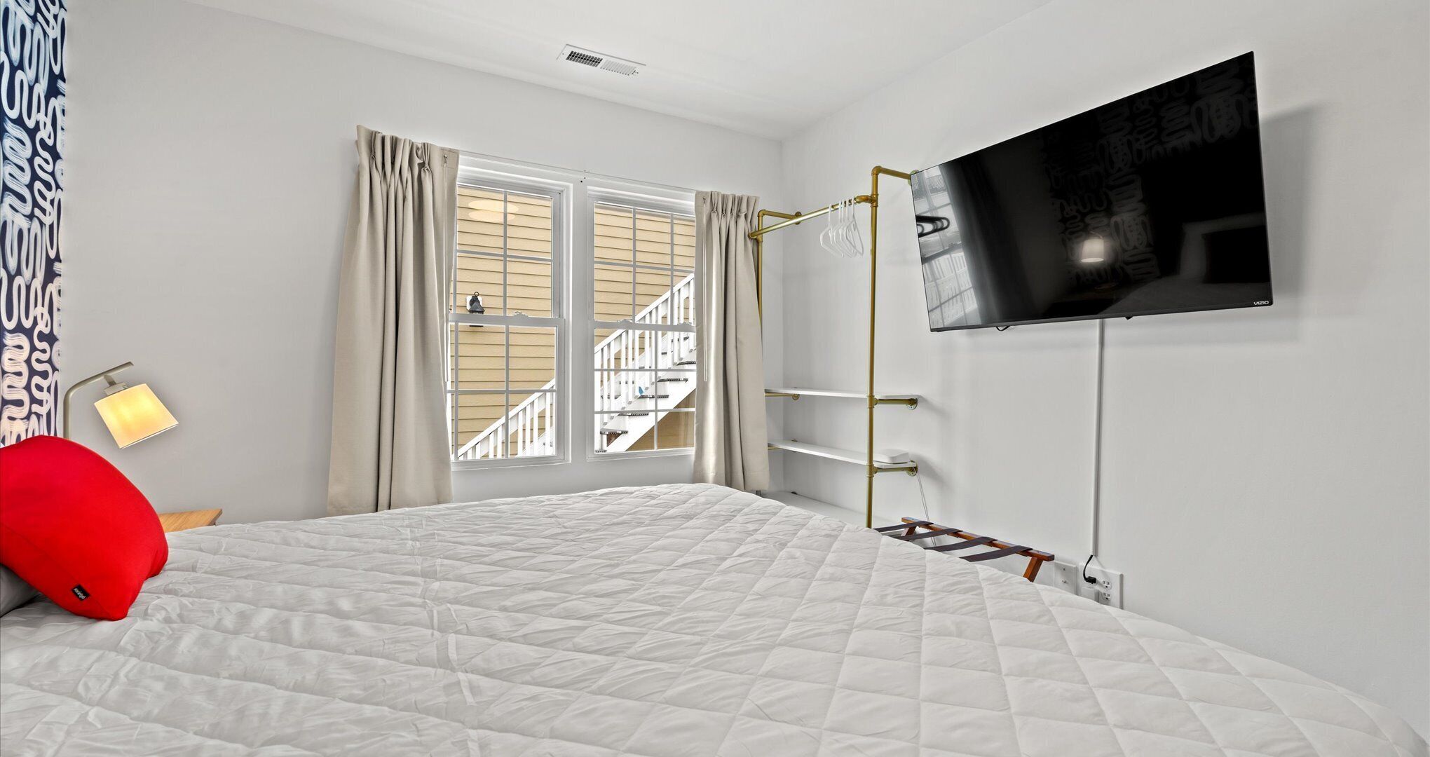 Een slaapkamer met een bed, een aan de muur bevestigde televisie, een raam met gordijnen en een uitzicht op een trap buiten.