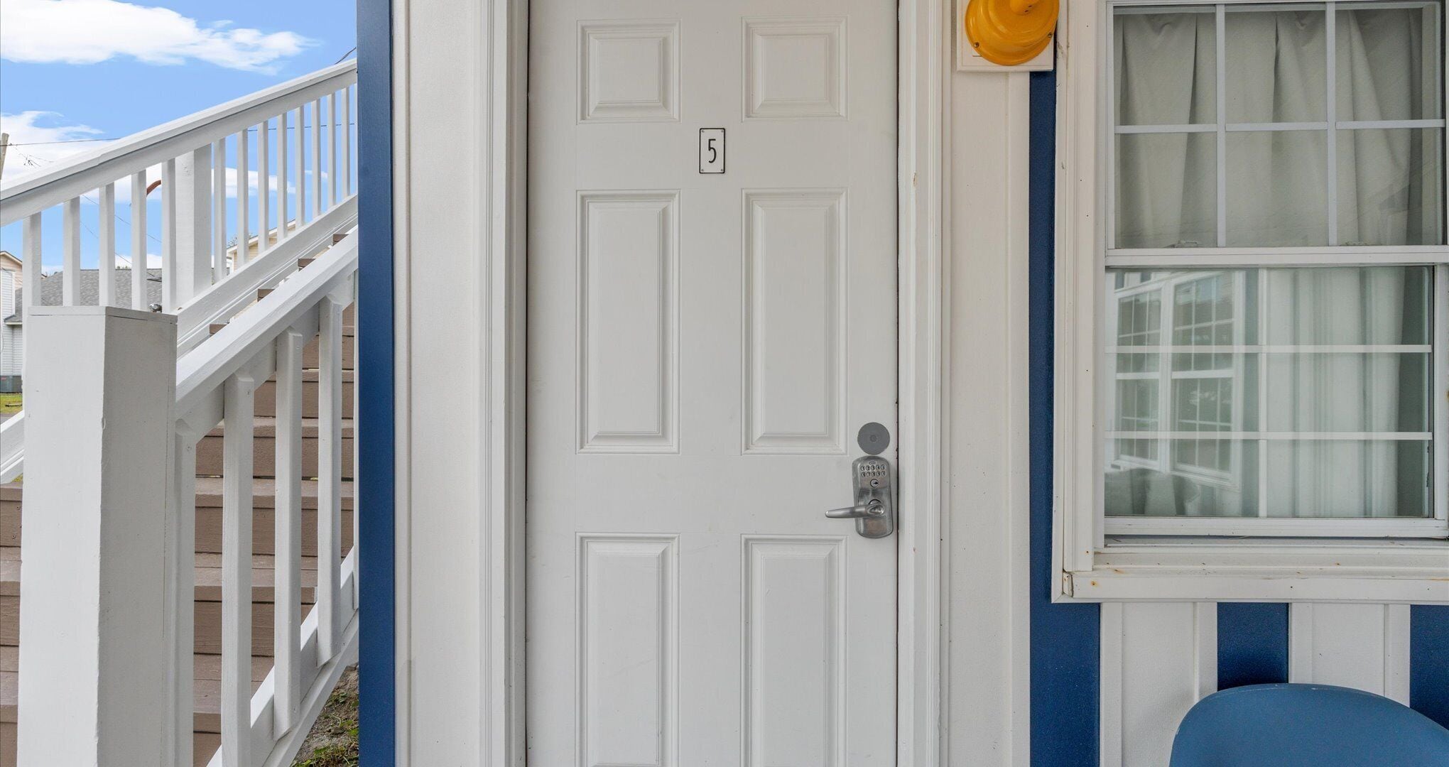 A white door with a number plate, a blue exterior wall, and a window with curtains.