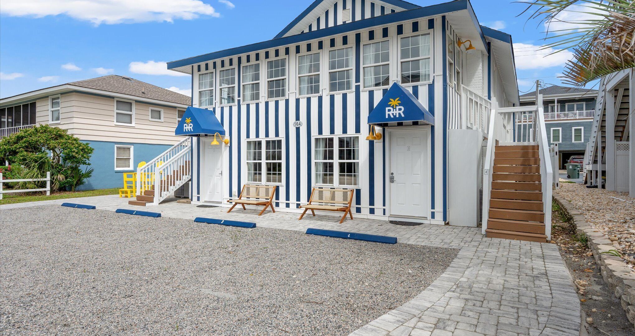 Un edificio con fachada azul y blanca a rayas, una entrada principal con puerta y dos bancos afuera.