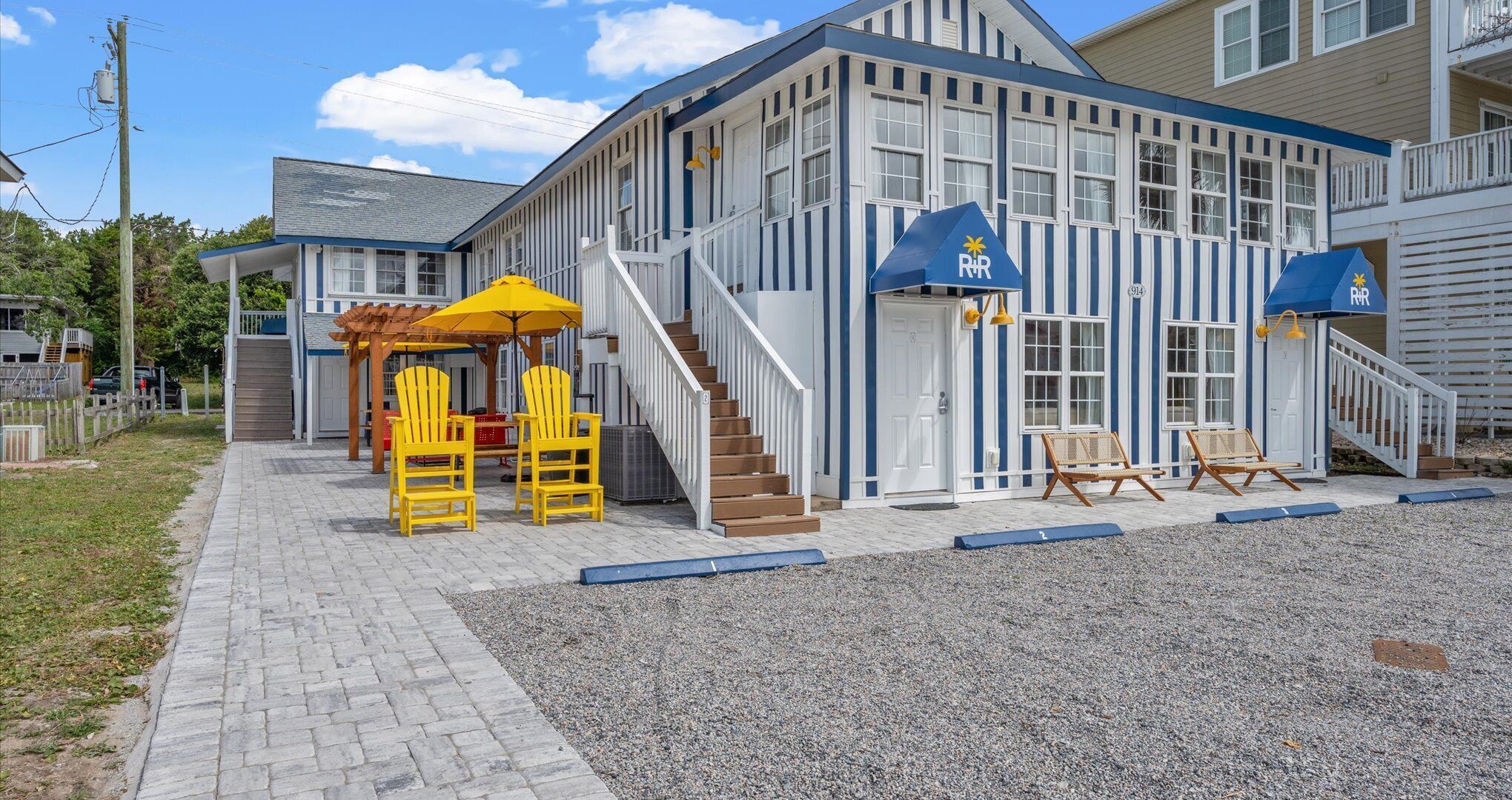 A building with a blue and white exterior, a covered outdoor seating area with yellow chairs, and a staircase leading to the entrance.