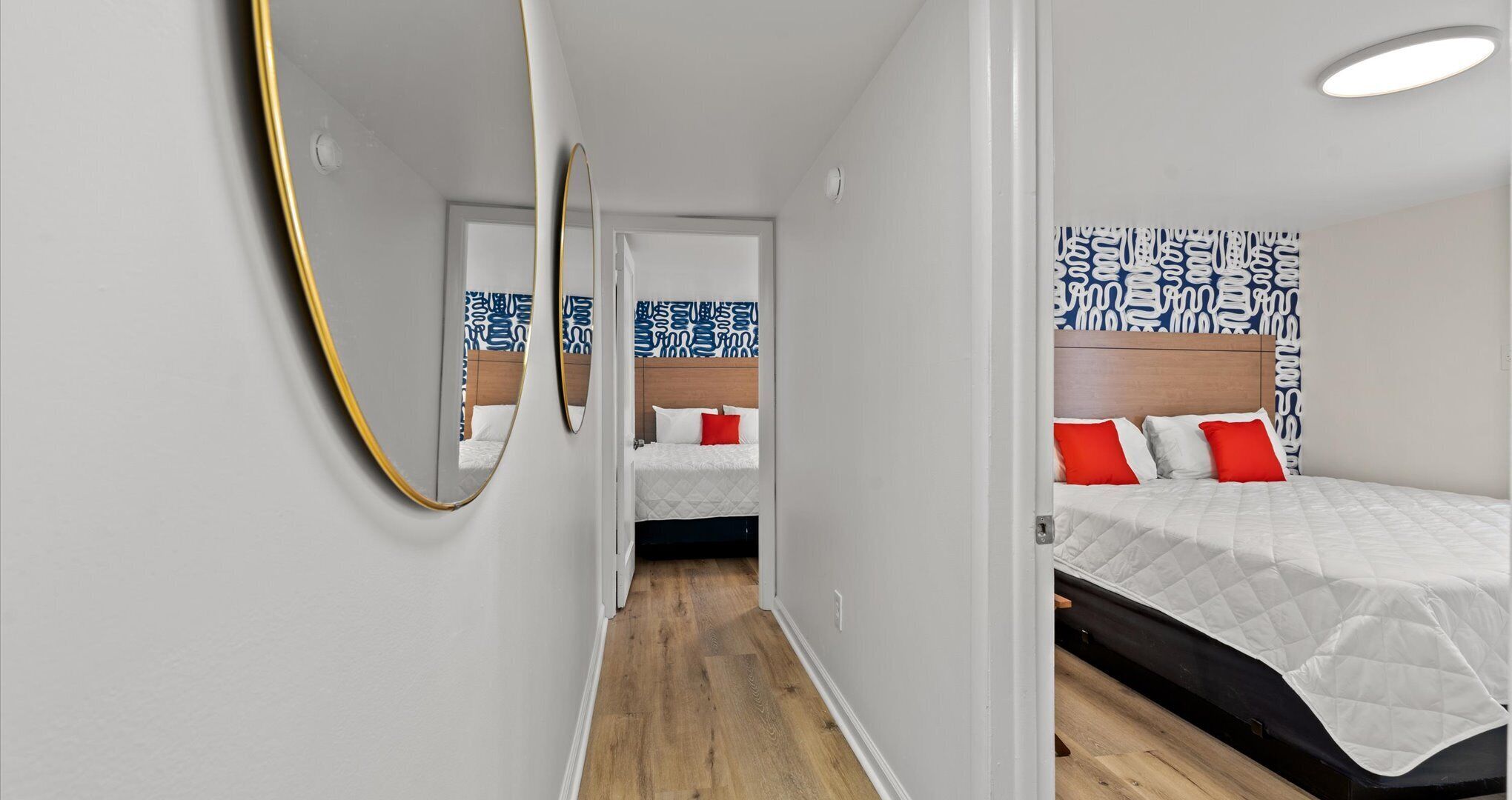 A narrow hallway leading to a bedroom with a bed, a mirror, and a patterned wall.