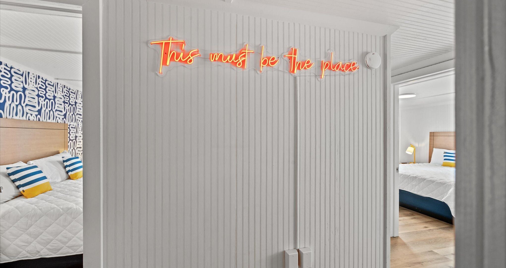 A hotel room with a striped wallpaper, a bed with blue and yellow striped pillows, and a neon sign on the door that reads "This must be the place."