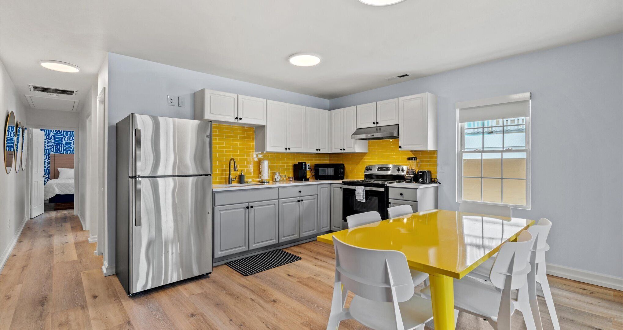A modern kitchen with white cabinets, a stainless steel refrigerator, a yellow-tiled backsplash, and a dining area with a yellow table and white chairs.