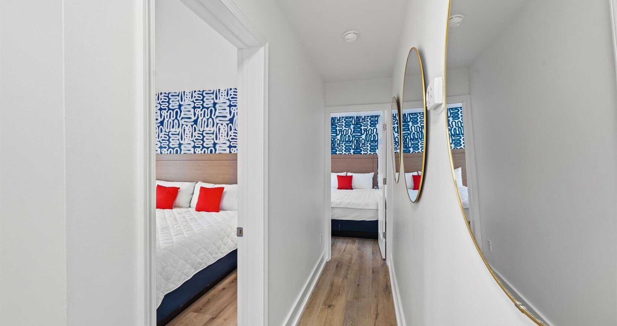 Ein schmaler Flur, der zu zwei Schlafzimmern führt, wobei jedes Zimmer ein Bett, rote Kissen und eine blau gemusterte Wand aufweist.