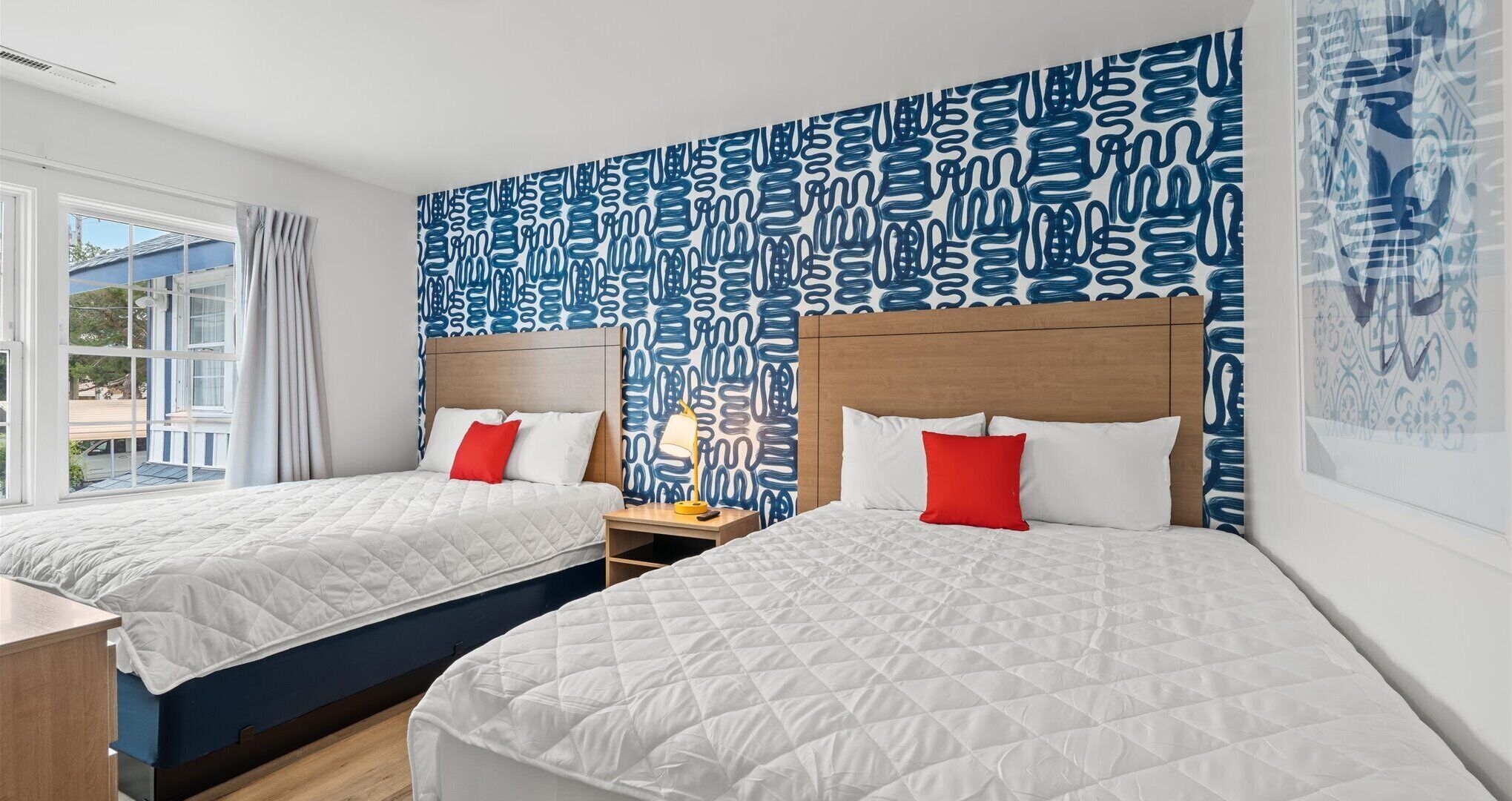 A hotel room with two beds, a wooden headboard, a nightstand with a lamp, and a window with curtains.