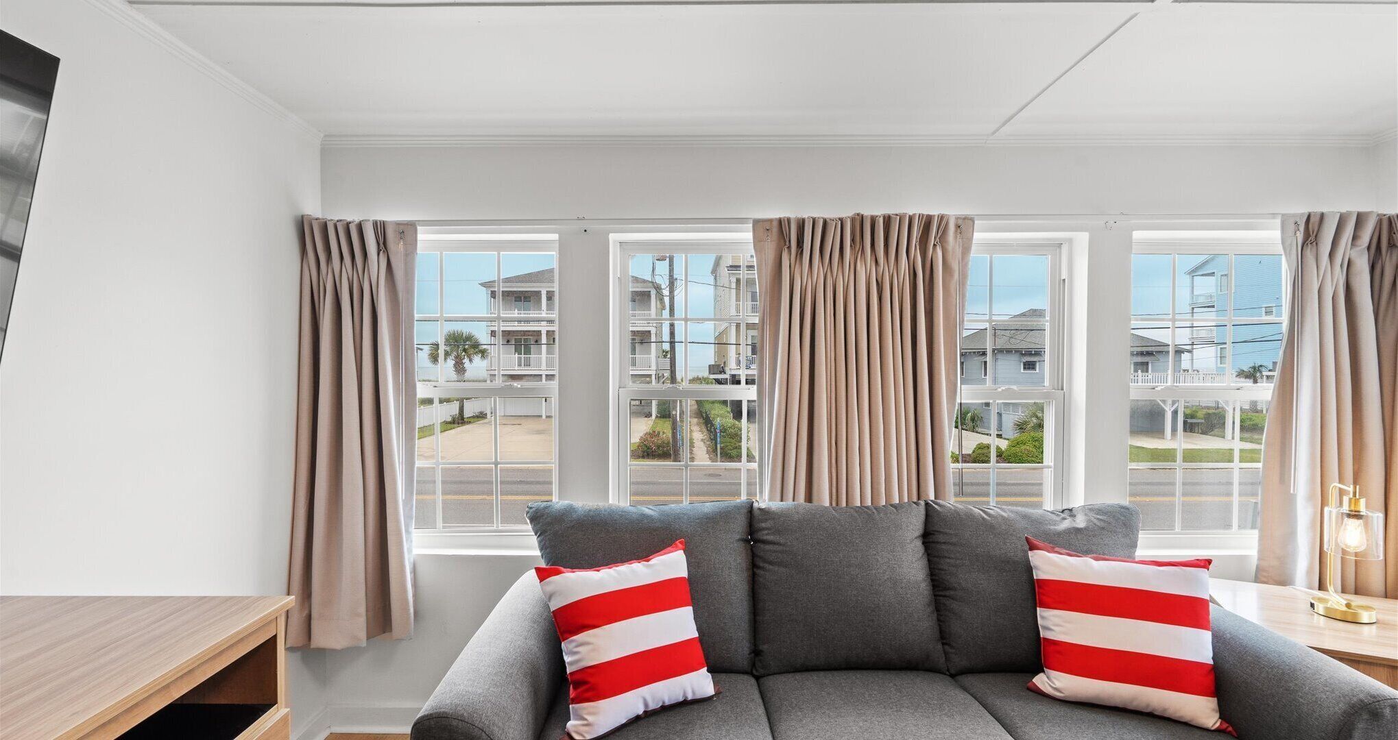 A modern living room with a grey sofa, beige curtains, and a view of a residential area through large windows.