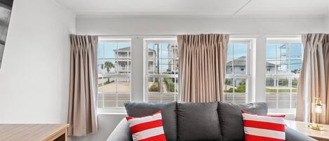 A modern living room with a grey sofa, beige curtains, and a view of a residential area through large windows.