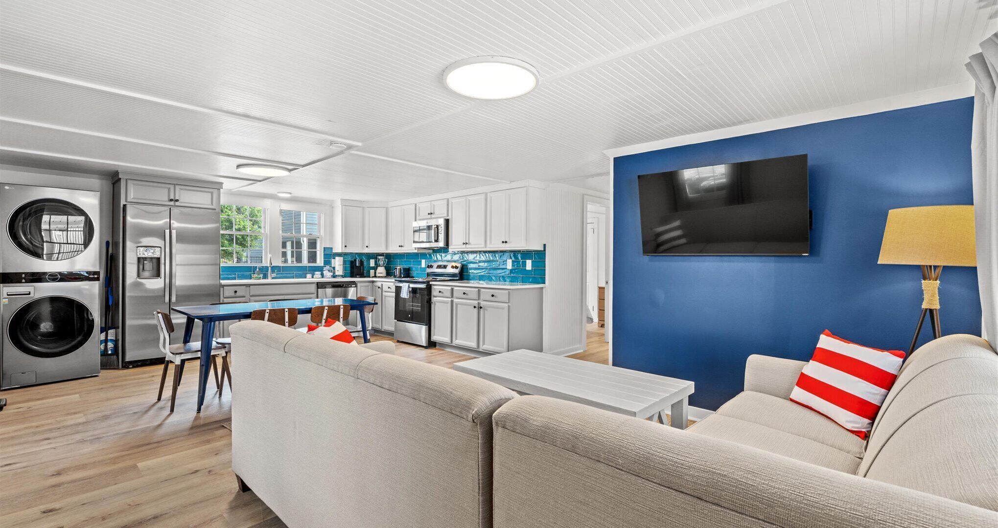 A modern kitchen with white cabinets, a blue backsplash, and stainless steel appliances. There is a dining area with a table and chairs, and a living room with a sofa and a TV mounted on a blue wall.