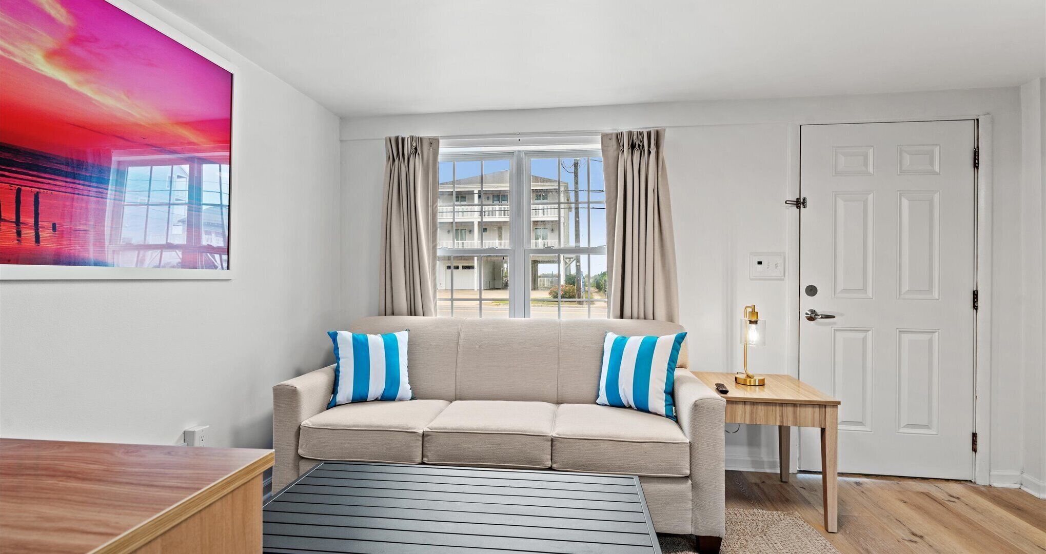 A room with a beige sofa, two striped pillows, a small wooden side table with a lamp, a large window with curtains, and a painting on the wall.