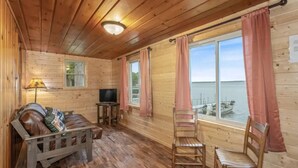 Eine Hütte mit Holzvertäfelung, einer Couch, einem Fernseher und Blick auf das Wasser.