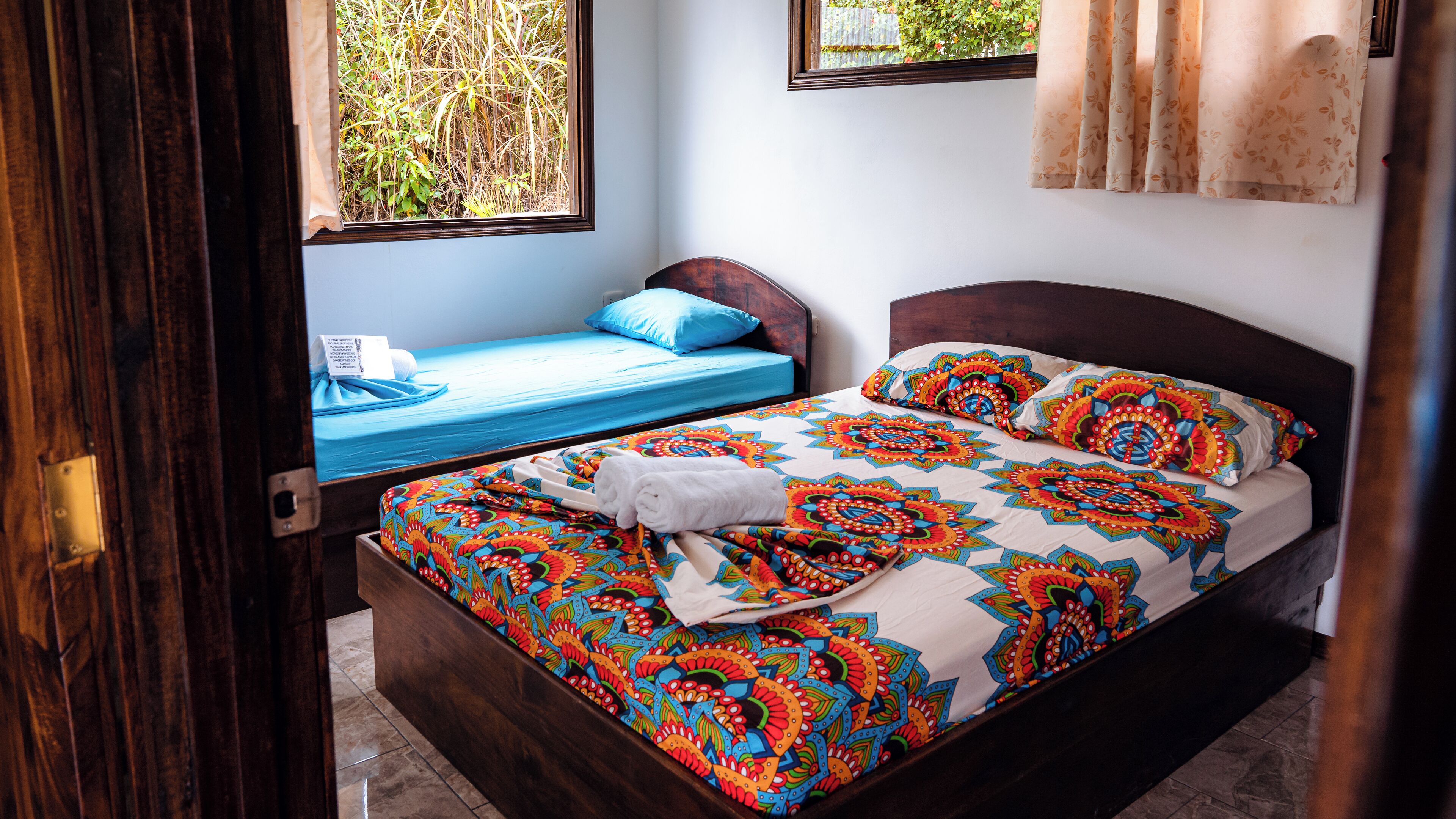 Una habitación con una cama de madera, una cama más pequeña con colchón azul y una ventana con vistas a zonas verdes.