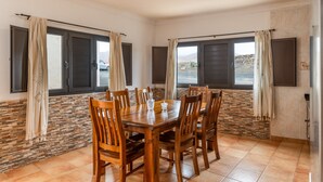 Un área de comedor con una mesa y sillas de madera, ventanas grandes con cortinas y una pared de piedra.