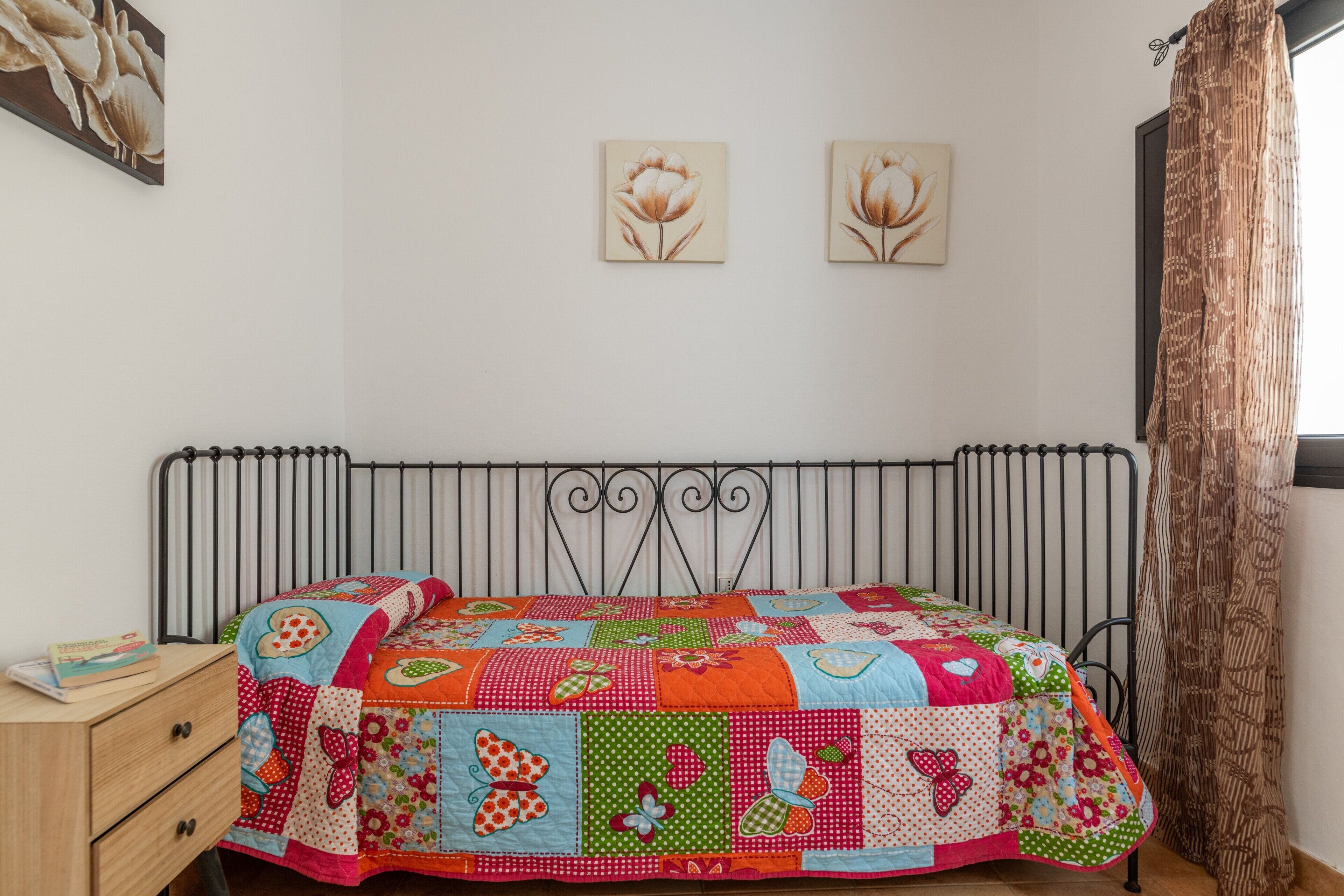 Une chambre à coucher avec un cadre de lit en métal, une courtepointe aux couleurs vives, une table de chevet en bois et deux tableaux représentant des fleurs, accrochés au mur.
