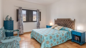 Un dormitorio con cama, mesita de noche, cómoda, silla y ventana con cortinas.